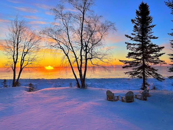 A snowy landscape with trees and a sunset over a body of water.