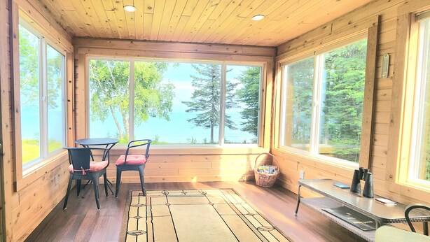 A wooden cabin with a large window overlooking a forest and a lake. Inside, there is a table with two chairs and a small rug.