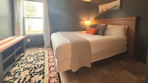 A bedroom with a bed, a nightstand, a lamp, a window with curtains, and a painting on the wall.