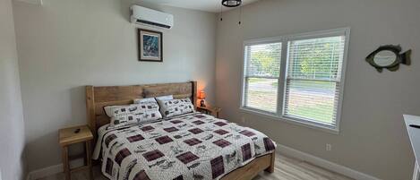 A bedroom with a wooden bed, a ceiling fan, an air conditioner, a window with blinds, and a framed picture on the wall.