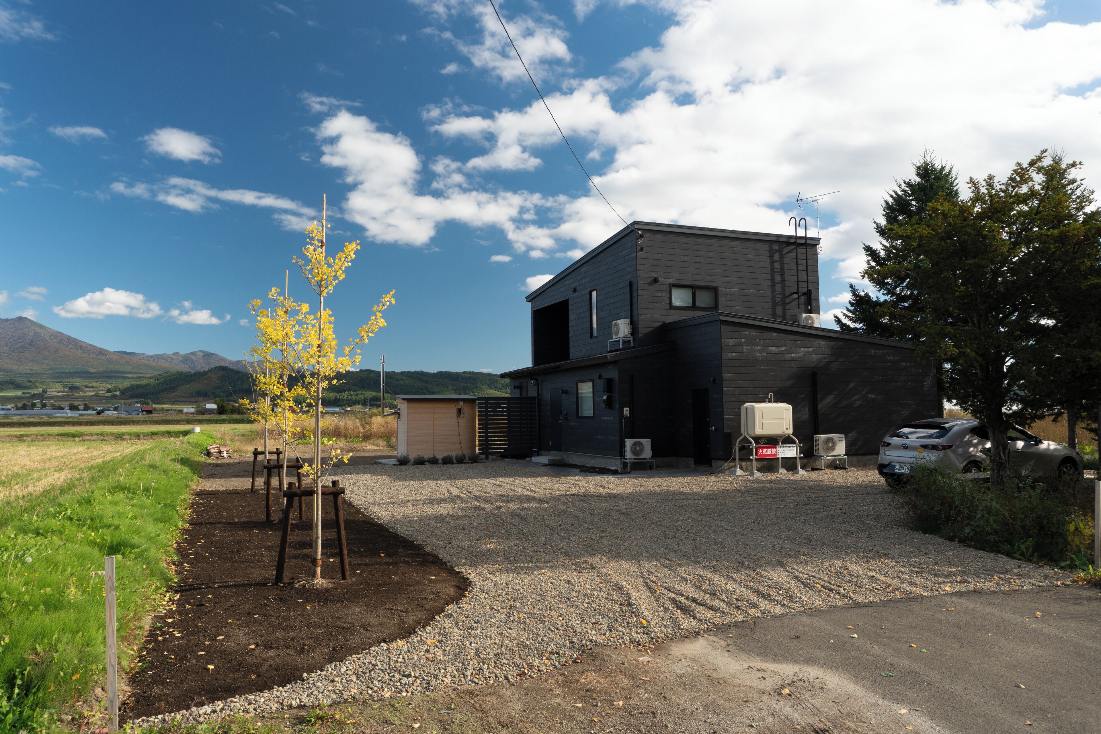 A modern two-story building with a gravel driveway, a small shed, and a tree with yellow leaves.