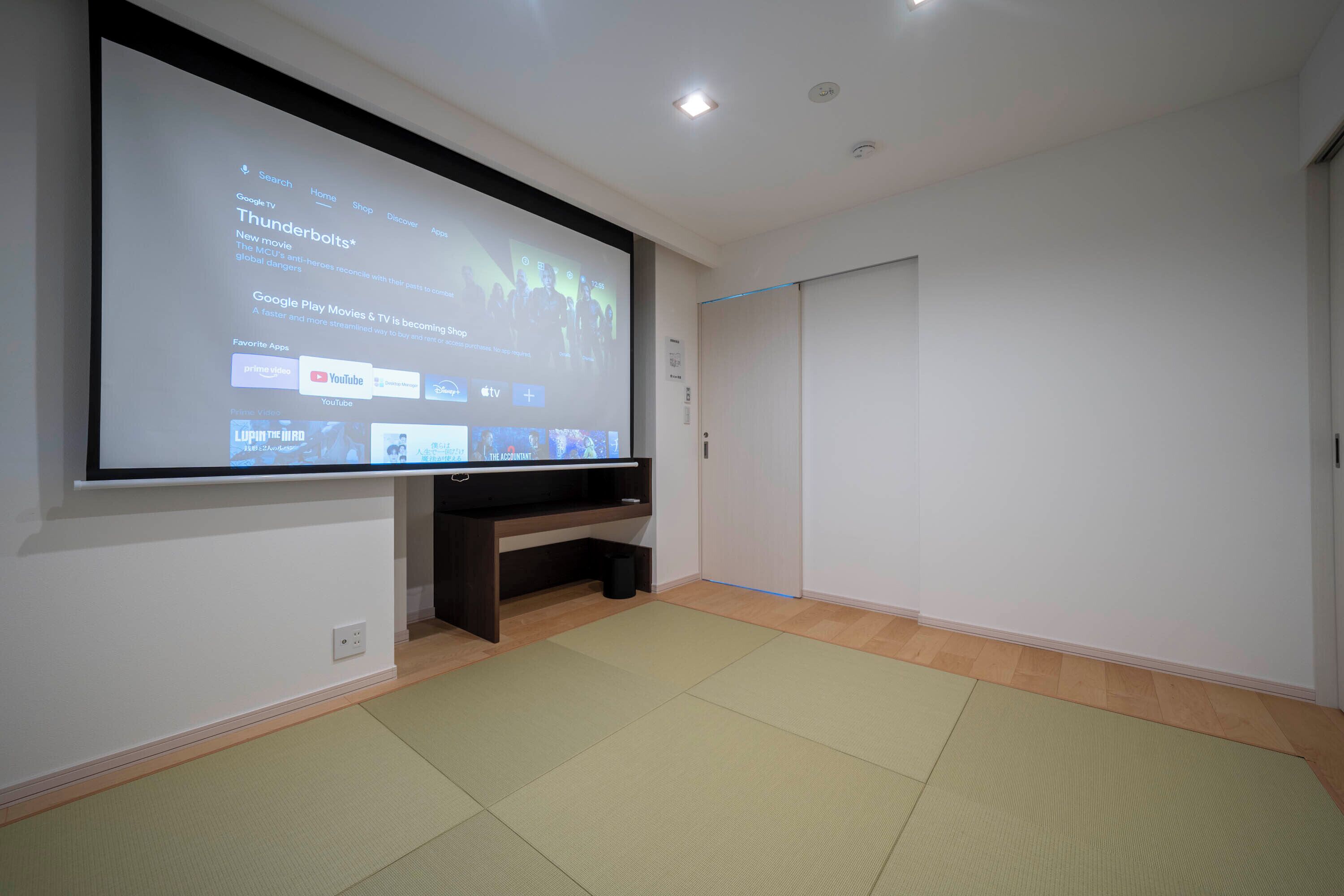 Ein Zimmer mit einem großen Flachbildfernseher an der Wand, einer dunklen Holzunterhaltungsecke darunter und einem Tatami-Boden.