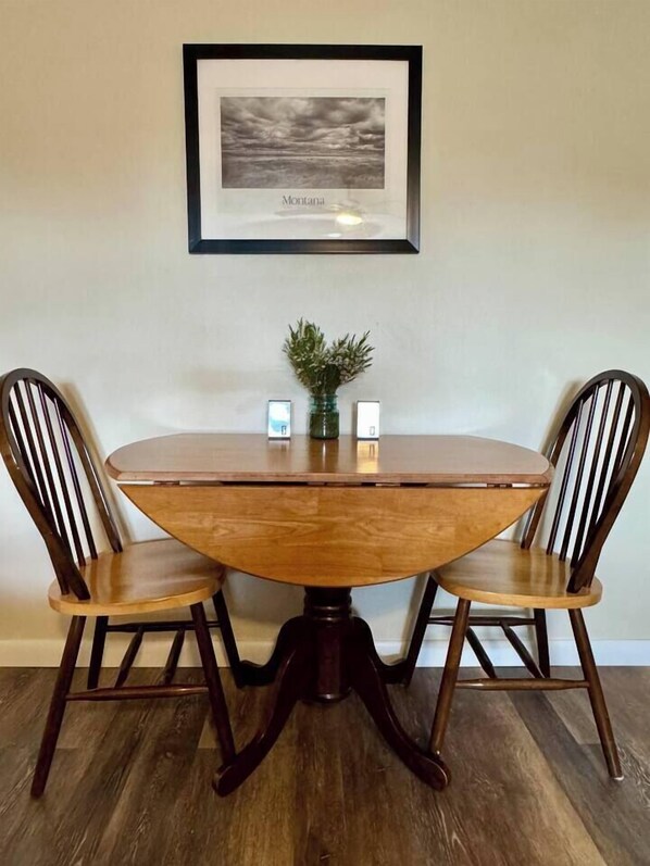 Une table de salle à manger ronde en bois, accompagnée de deux chaises, un tableau encadré accroché au mur et une petite plante posée sur la table.