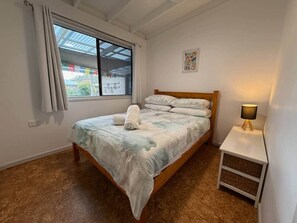 Een slaapkamer met een bed, nachtkastje met lamp, raam met gordijnen en een wanddecoratie.