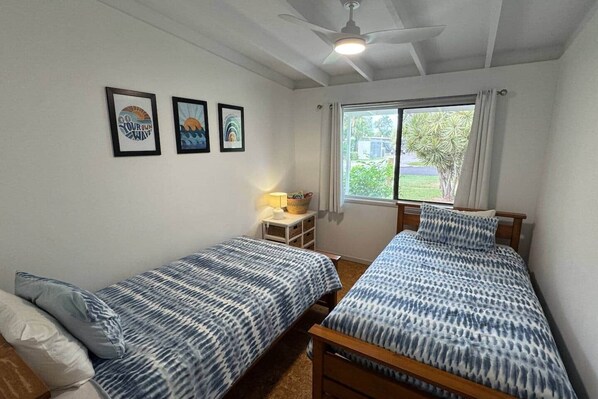 Une chambre avec deux lits, un ventilateur de plafond, une fenêtre avec des rideaux et trois tableaux encadrés accrochés au mur.