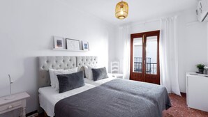 Een slaapkamer met een gestoffeerd hoofdeinde, een bed met grijs en wit beddengoed, een nachtkastje, een hanglamp, een raam met witte gordijnen en een balkon met een hekwerk.