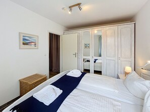 Una habitación de hotel con cama, armario, mesita de noche con lámpara y un pequeño taburete de madera.