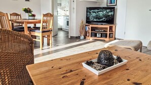 A modern living room with a wooden dining table, wicker chairs, a TV on a wooden stand, and a kitchen in the background. - Beautiful apartment for 4 people with WIFI, TV and terrace (Wolgast)