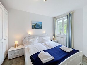 Une chambre à coucher avec un lit, des tables de chevet, une fenêtre avec des rideaux, un tableau au mur et un radiateur.