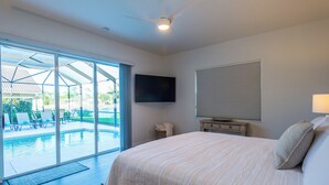 Un dormitorio con una cama grande, un televisor montado en la pared, un ventilador de techo y un ventanal corredero que da a una zona de piscina.