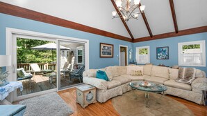A living room with a large sectional sofa, a glass coffee table, and a chandelier. There is a sliding glass door leading to an outdoor patio with chairs and a table. - Cape Cod Escape – 1 Mile to Beach, A/C & Grill (Centerville)