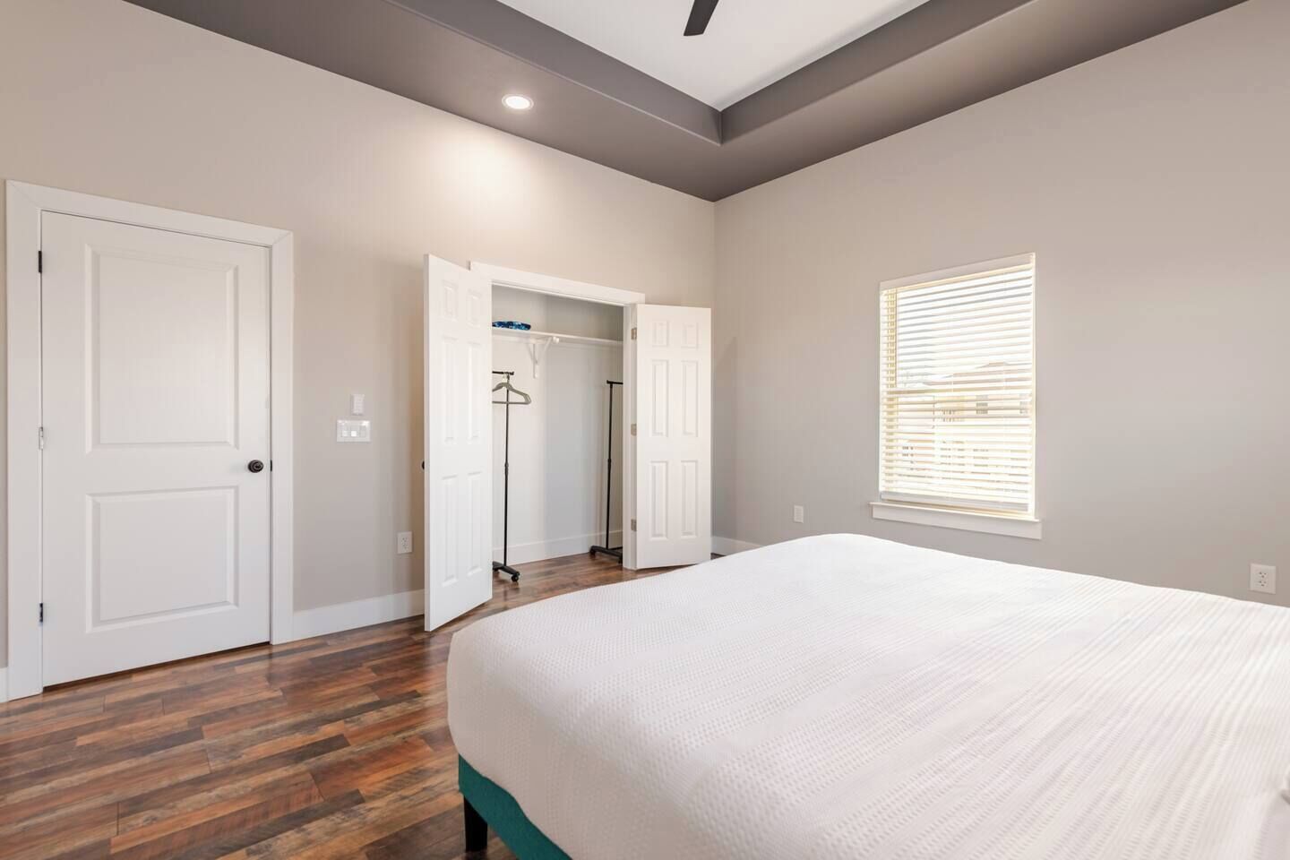 A bedroom with a large bed, wooden flooring, a closet with hanging clothes, and a window with blinds.
