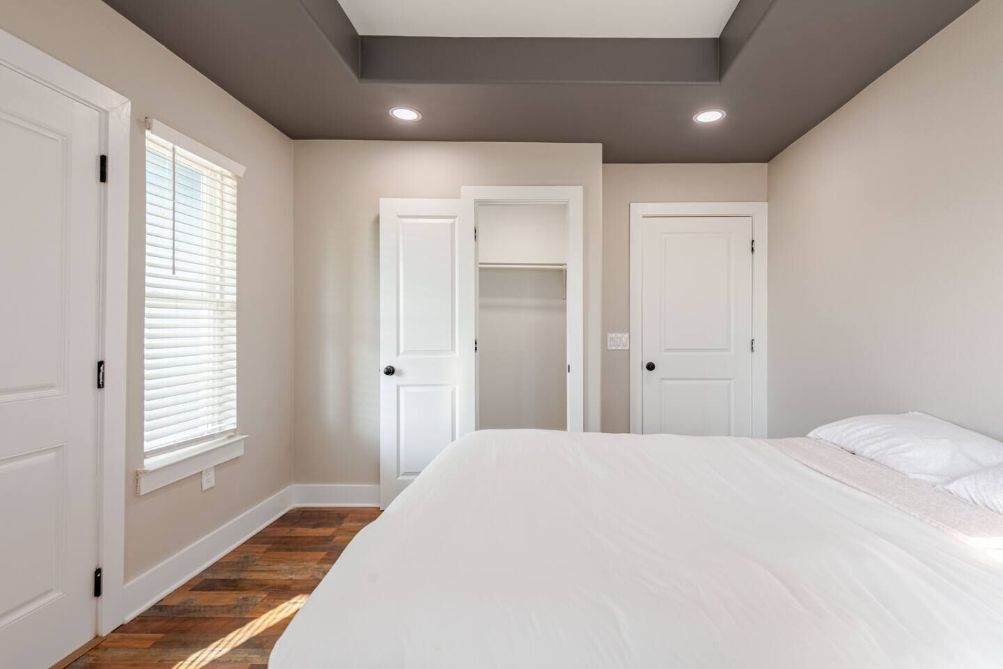 A bedroom with a large bed, two white doors, a closet, and a window with blinds.