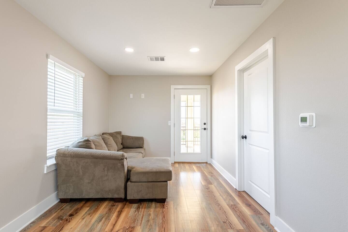 A room with a sectional sofa, a door with a glass panel, and a thermostat on the wall.