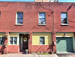 A red brick two-story building with a green garage door, a front door with a hanging flower basket, and a small front yard with potted plants.