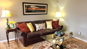 Un salon avec un canapé en cuir marron, une table basse en verre, un tapis à motifs, deux lampes et un tableau encadré accroché au mur.