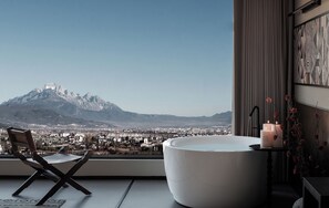 Un baño moderno con una bañera grande, una silla y vistas a una montaña y un paisaje urbano.