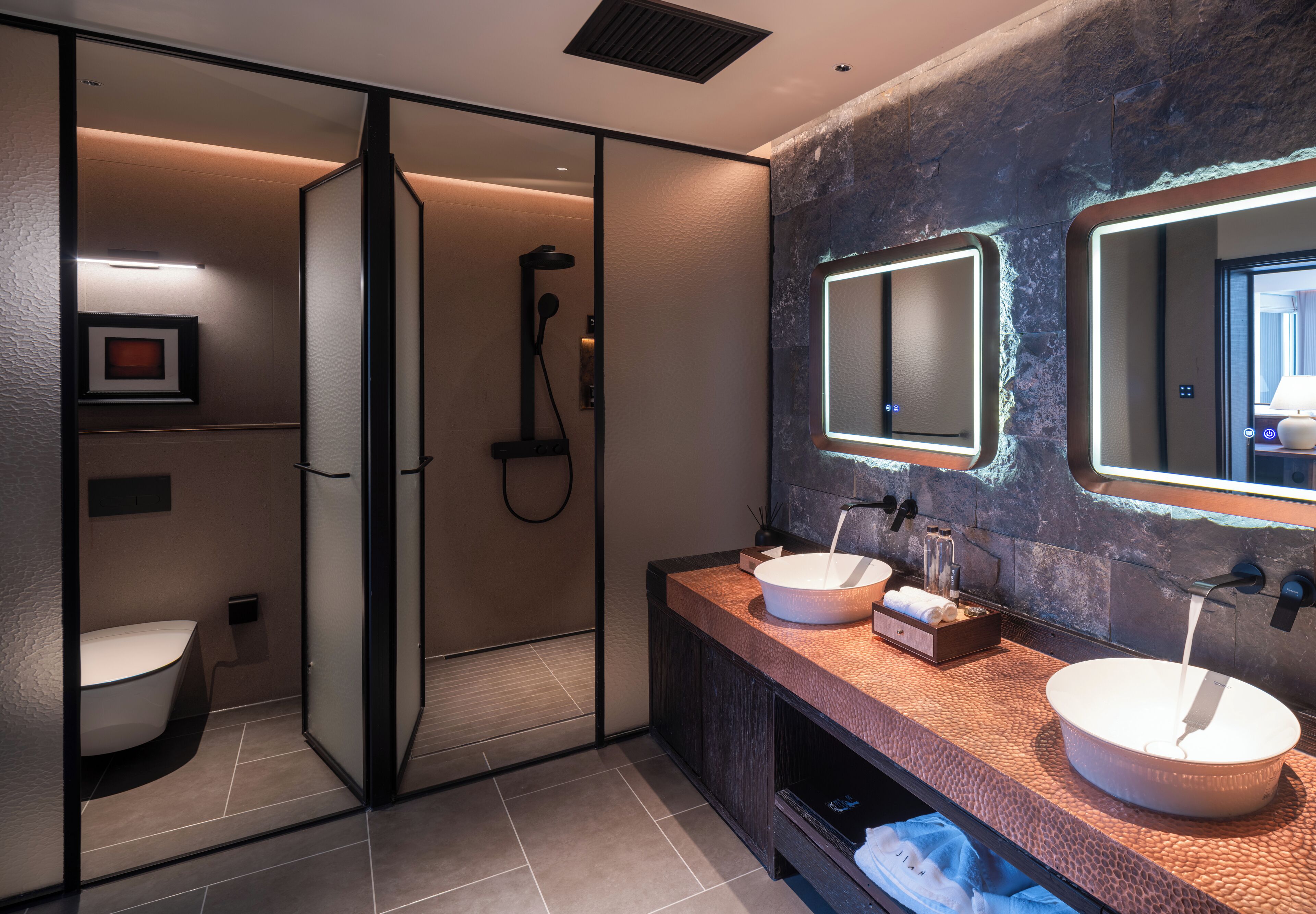 Un baño moderno con ducha envuelta en vidrio, dos lavamanos con espejos redondos y una pared con textura.