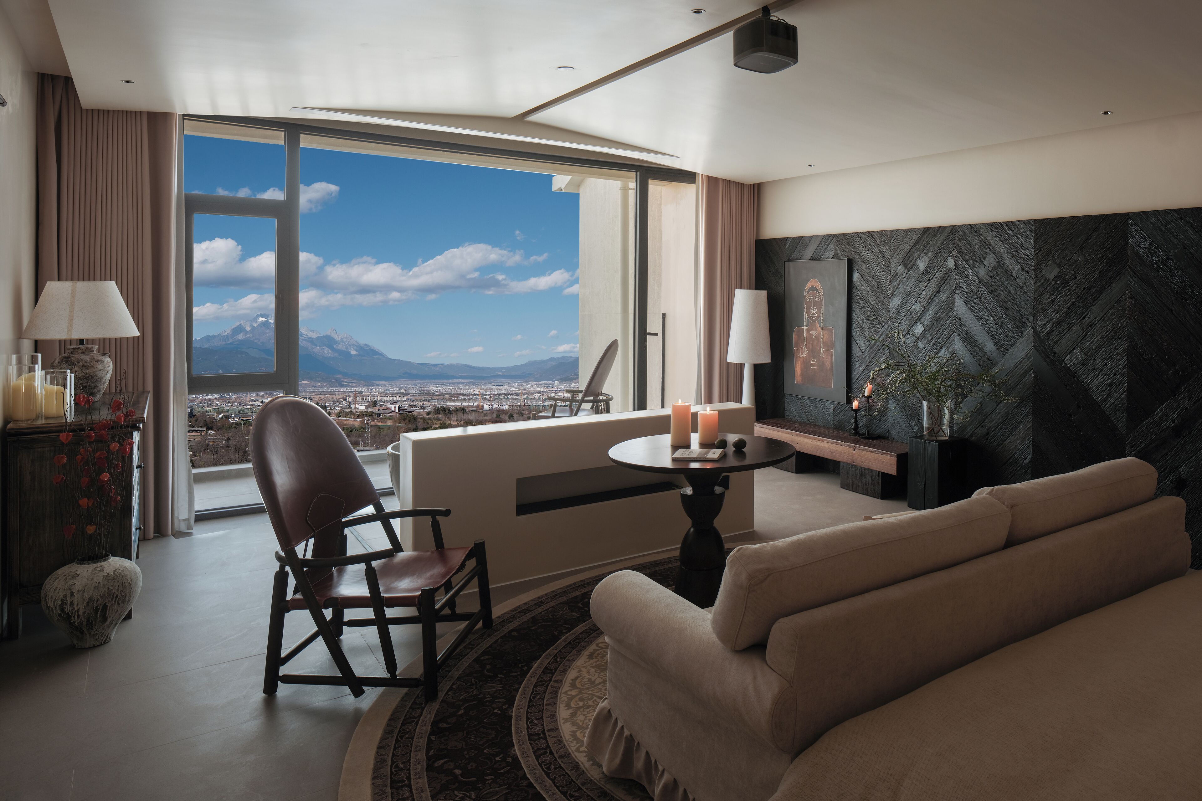 A modern hotel room with a large window offering a city view, a comfortable sofa, a small round table with candles, and a decorative vase.