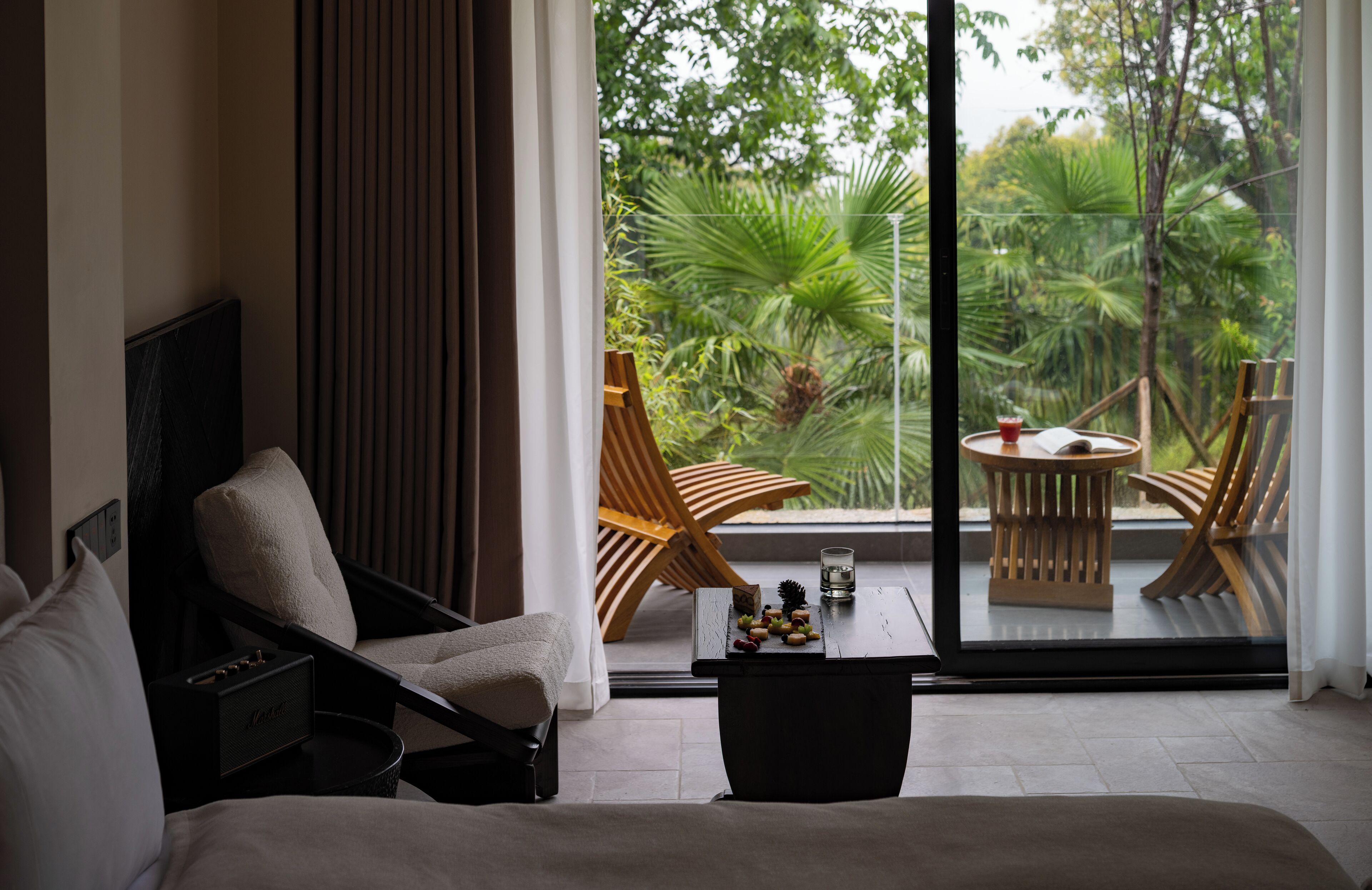 A modern room with a sofa, a wooden chair, a small table with fruits and a book, and a view of lush greenery through large windows.