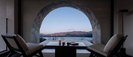 Una habitación con ventana abovedada de piedra que da a un lago y montañas. Hay dos sillas y una mesa con velas y libros.
