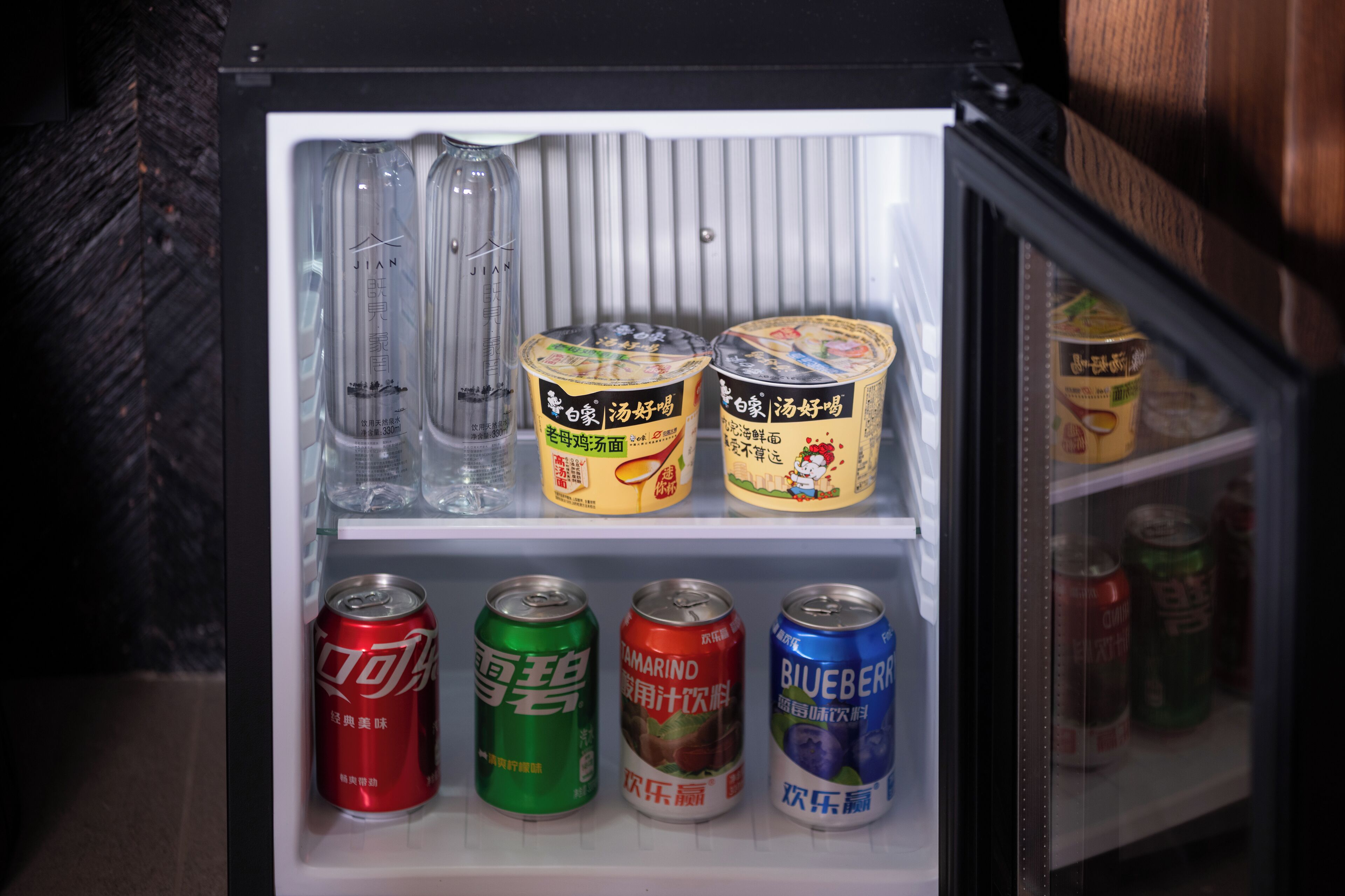 Un mini refrigerador con diversas bebidas, incluyendo latas de Coca-Cola, Sprite y Mountain Dew, así como dos recipientes de una bebida amarilla con texto en chino.