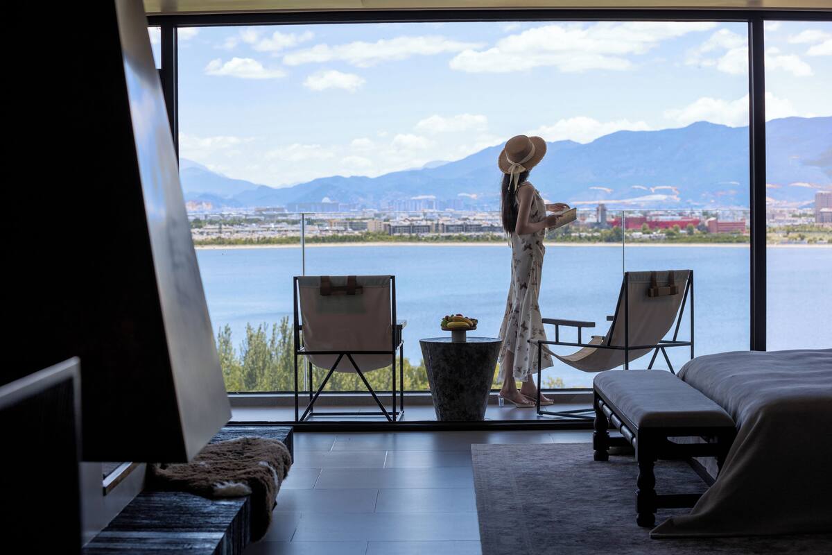 Una habitación con un ventanal grande que da a un cuerpo de agua y montañas, una cama, dos sillas y una mesita.