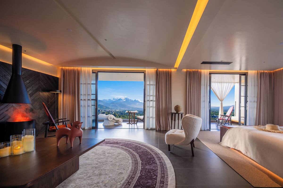 Una habitación de hotel moderna con una cama grande, un sofá, una silla, una chimenea y vistas a las montañas.