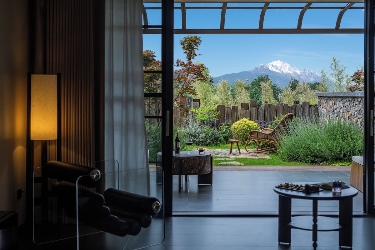 Un interior moderno con un ventanal grande, un sofá, una mesa y vistas a un jardín con cerca de madera y una montaña al fondo.