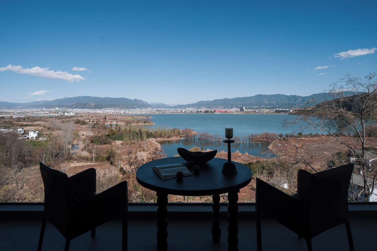 Una mesa con un libro y una vela, con vista a un lago y un paisaje urbano distante.