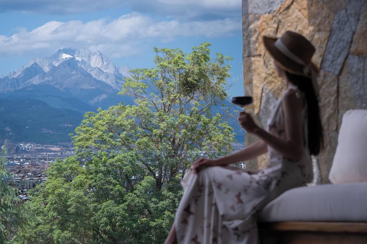 Una habitación con vistas a una cordillera, un árbol y una persona sentada junto a la ventana.