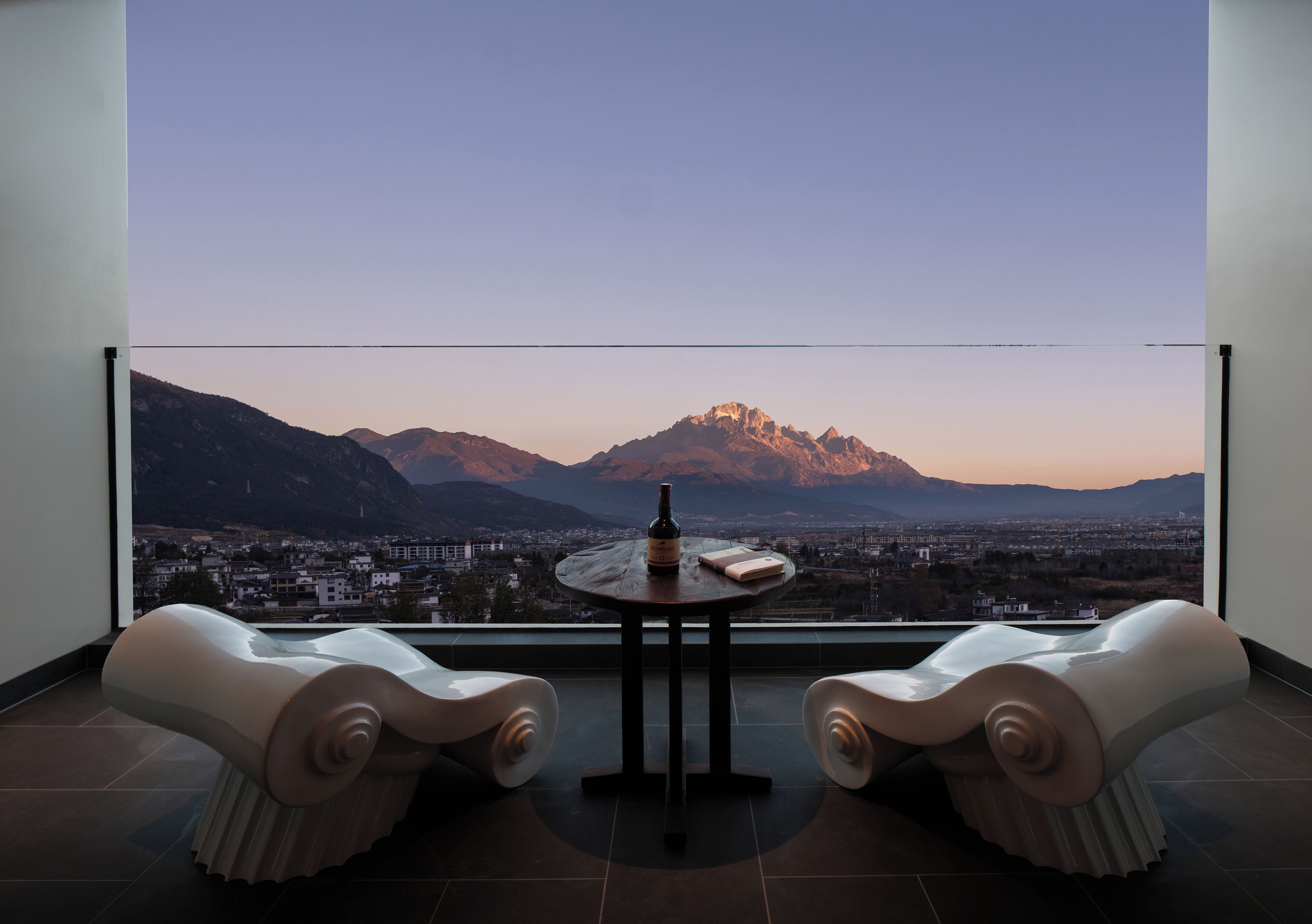A modern room with a panoramic mountain view, a round table with a bottle and book, and two sculptural chairs.