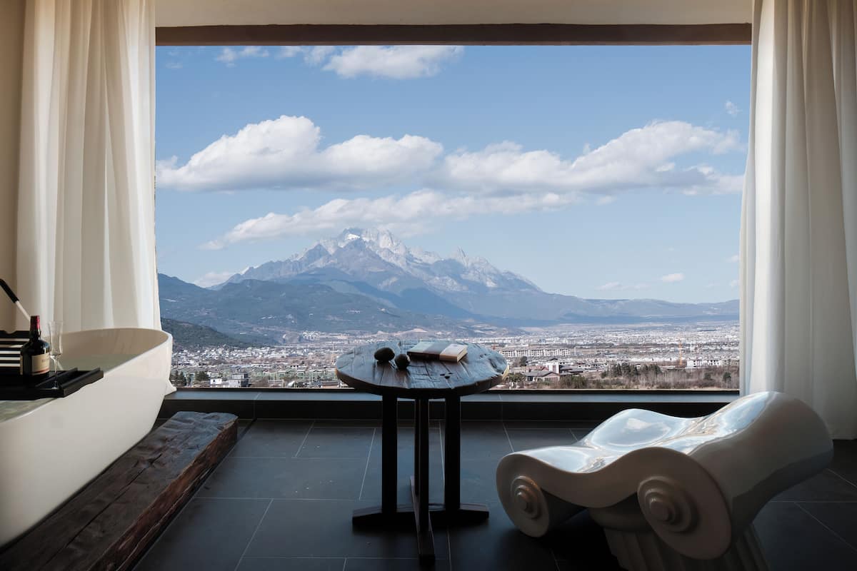Una habitación con vistas a la montaña, una mesa redonda con una botella y vasos, y una silla blanca.
