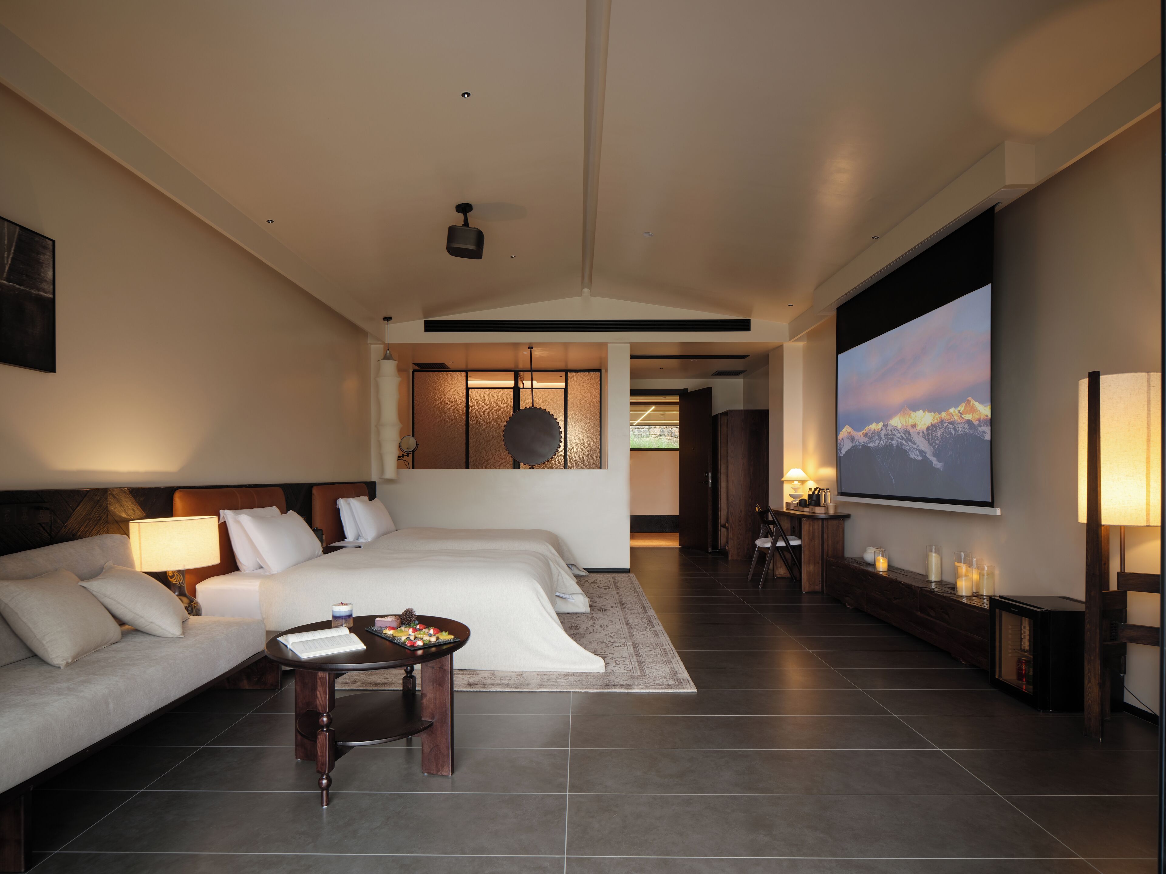 A modern hotel room with a large bed, a sofa, a small table with a laptop, and a TV displaying a mountain scene.