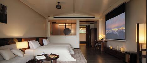 A modern hotel room with a large bed, a sofa, a small table with a laptop, and a TV displaying a mountain scene.