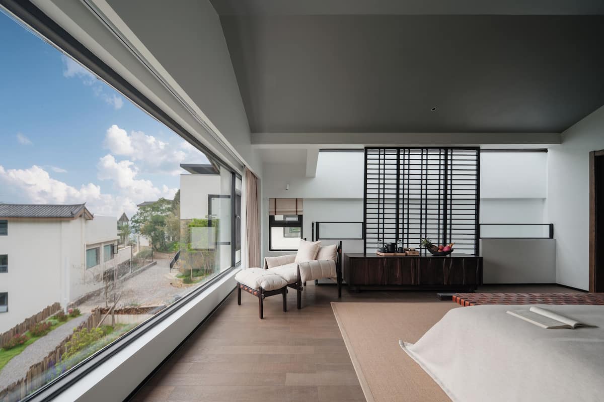 Un dormitorio moderno con un ventanal grande, una cama, una silla, una mesita y un biombo decorativo.