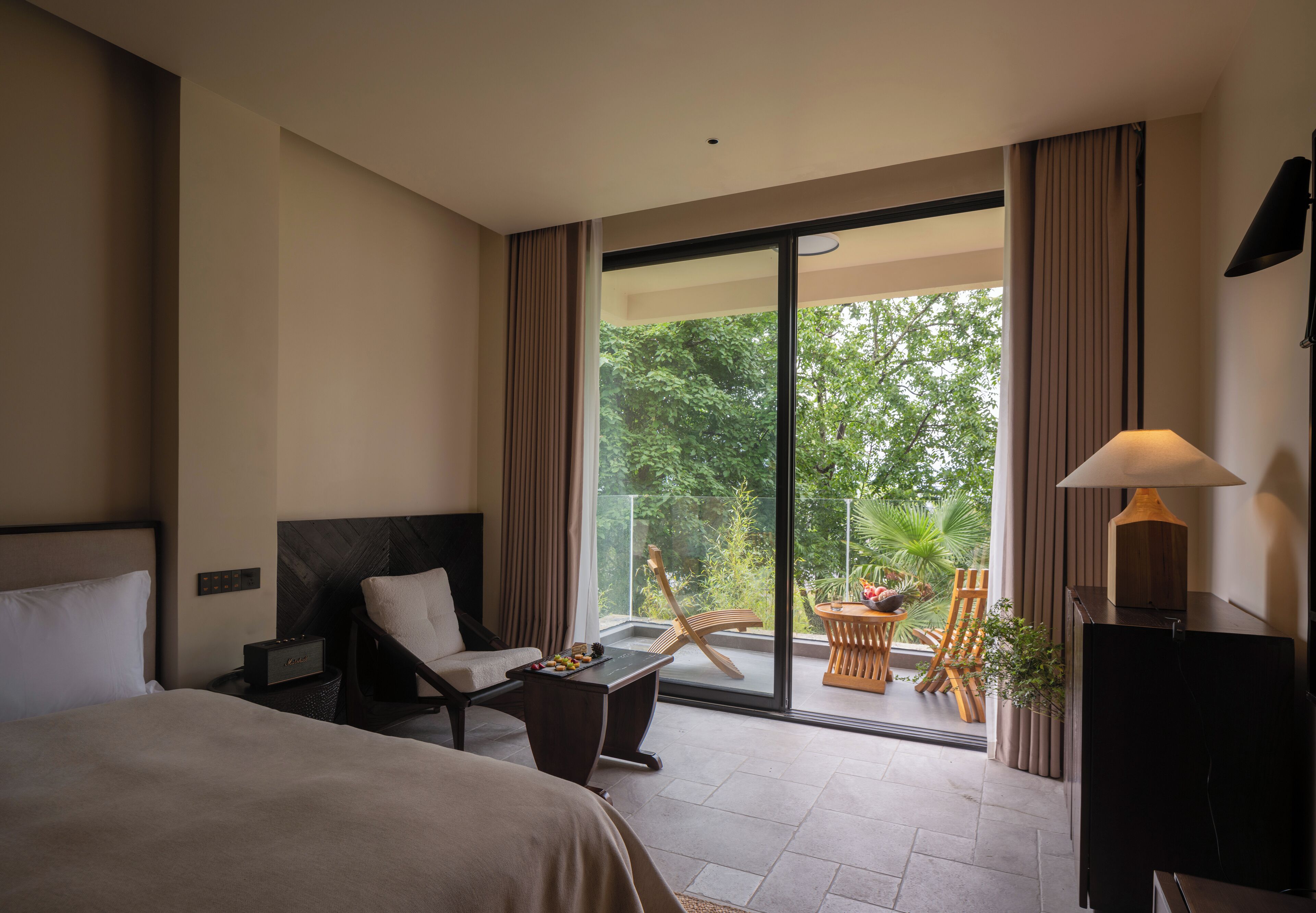 A hotel room with a bed, a desk, a chair, a table with a bowl of fruit, and a view of greenery through a sliding glass door.