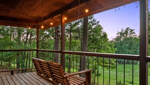 A wooden deck with a bench, hanging lights, and a view of a forest.