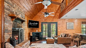 A cozy cabin interior with a fireplace, leather seating, a television, and a wooden ceiling.