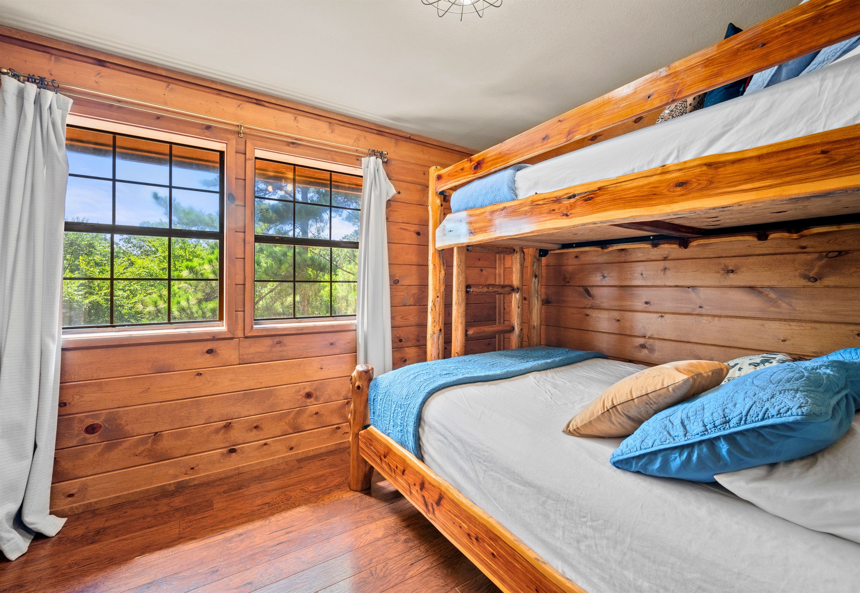 A bunk bed room with wooden walls, a window with curtains, and a view of greenery outside.