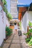 A narrow pathway with a suitcase, surrounded by greenery and a building with a blue roof.