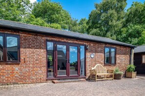 A brick building with large glass doors and windows, a wooden bench, and a potted plant. - Rural Farm Cottage | Outside Space | 2bds & 2baths (Stelling Minnis)