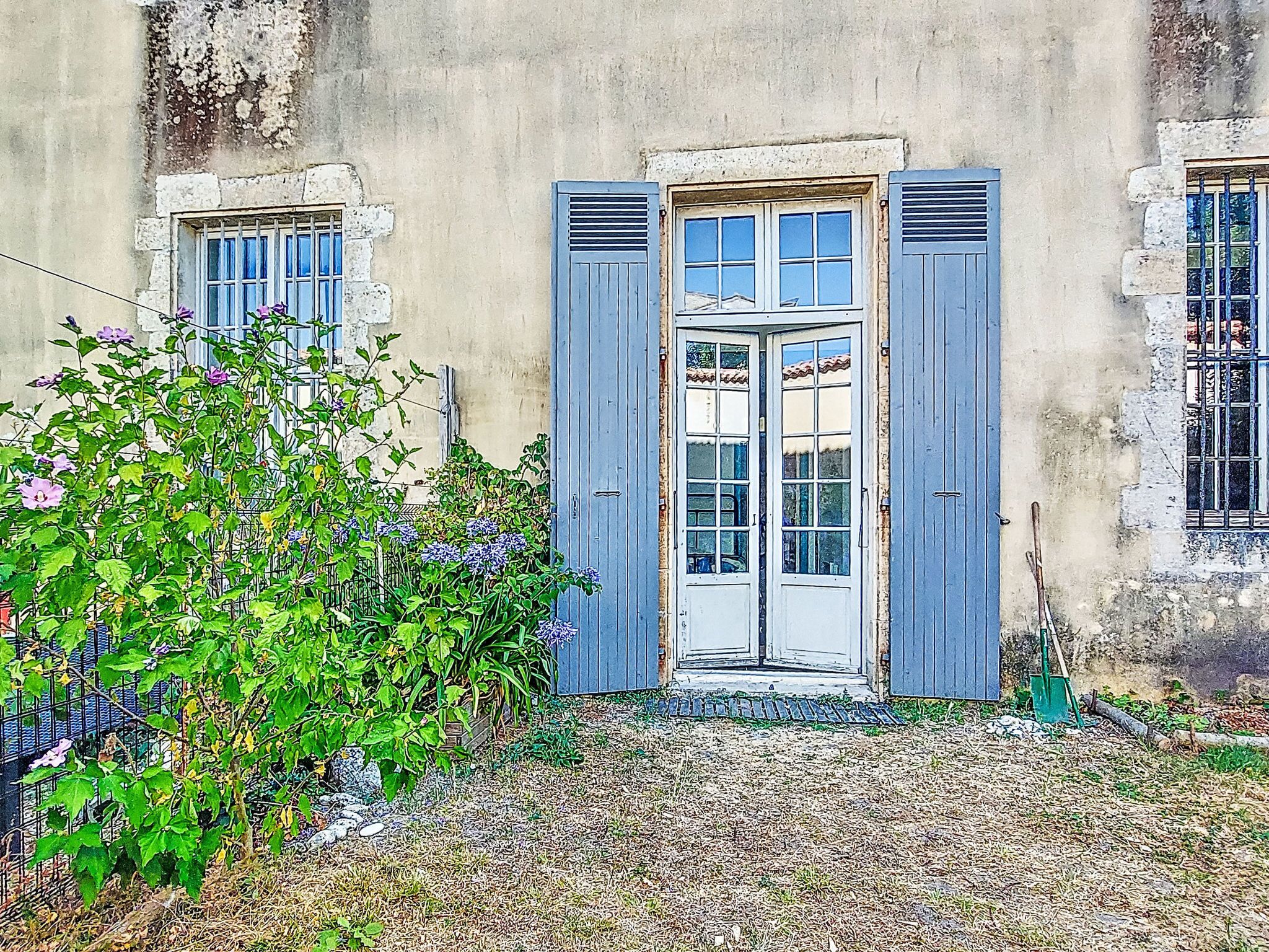A building with a central door flanked by two windows, a small garden area with plants, and a gravel path leading to the door.
