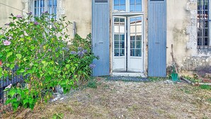 Un bâtiment doté d’une porte centrale, de part et d’autre de laquelle se trouvent deux fenêtres, ainsi qu’un petit jardin avec des plantes et un chemin de gravier menant à la porte.