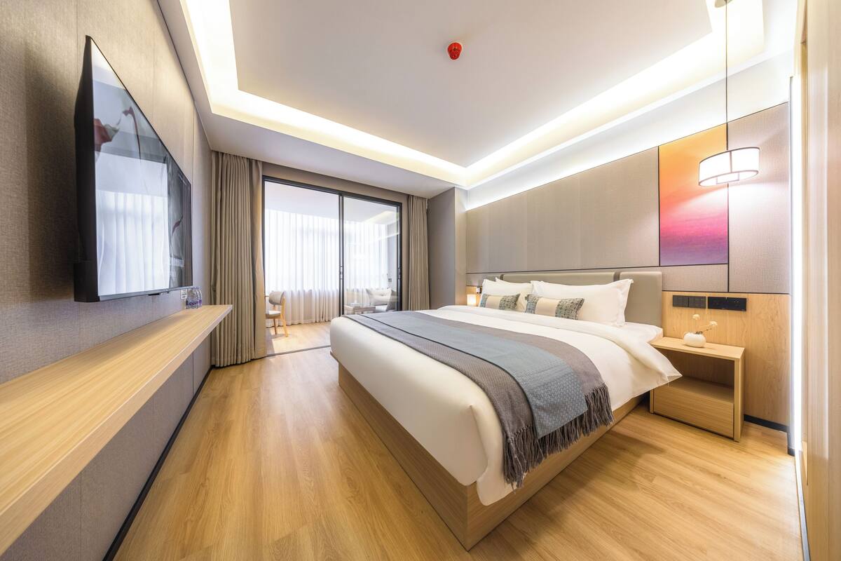 A modern hotel room with a large bed, a flat-screen TV, a wooden nightstand, and a large window with curtains.