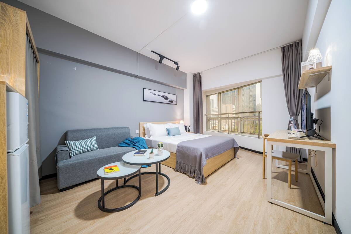 A modern hotel room with a grey sofa, a single bed, a small round table, a desk, and a large window offering a city view.
