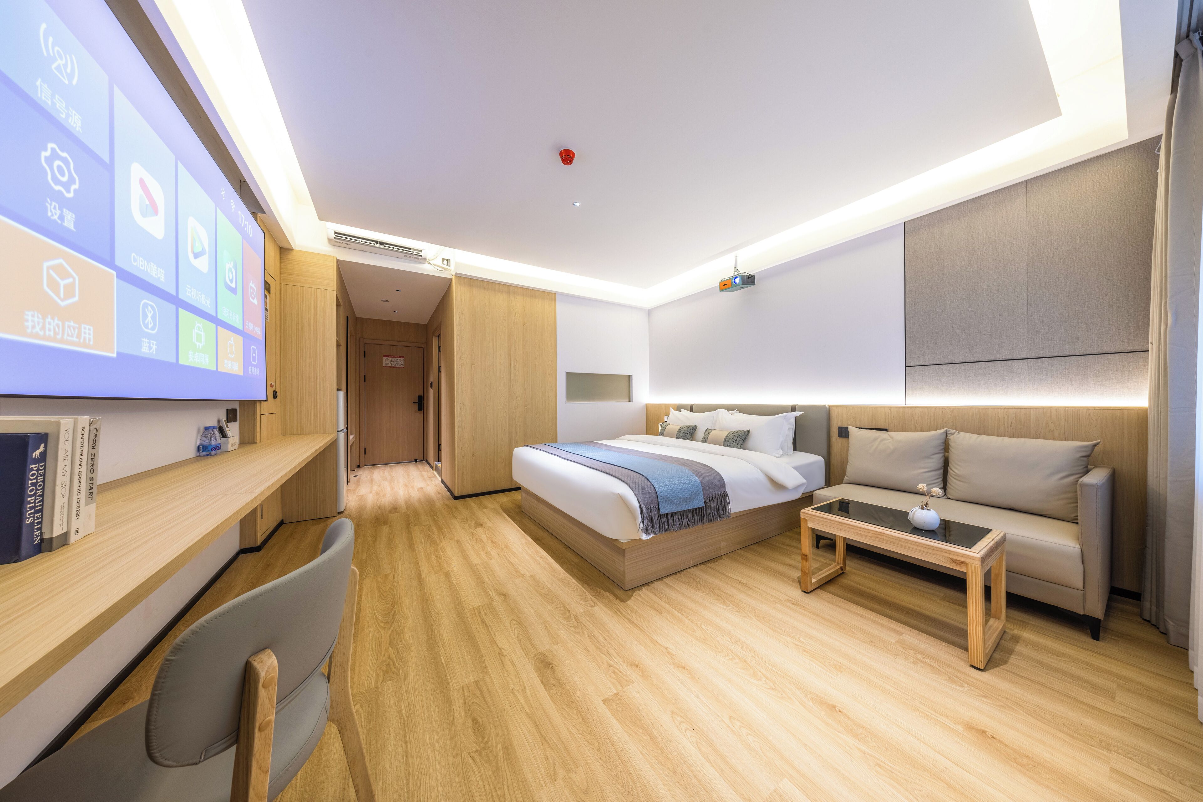 Habitación de hotel moderna con piso de madera, una cama con cubrecama azul y blanco, un sofá, un escritorio con silla y una pantalla grande que muestra iconos.