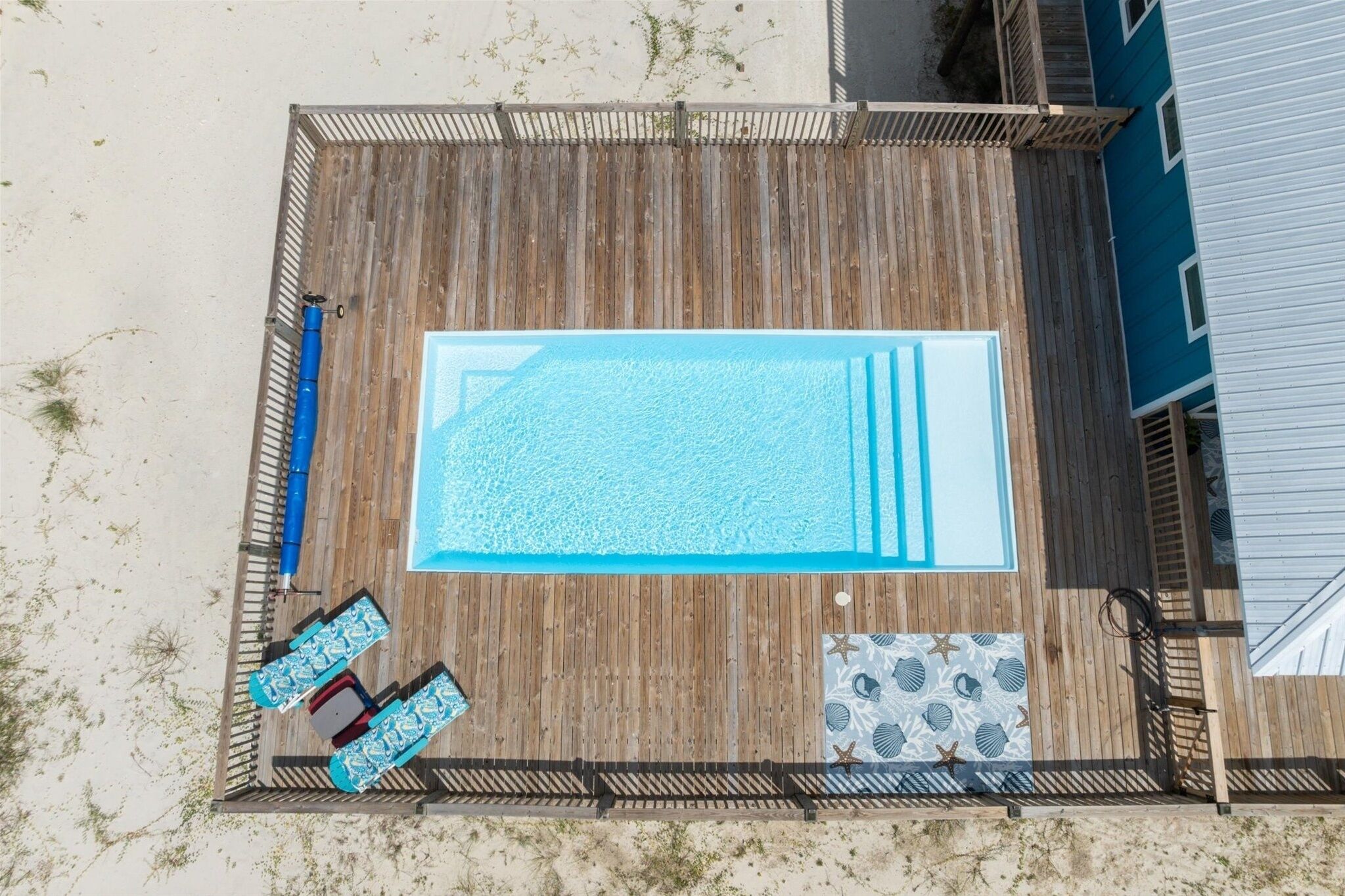 An aerial view of a wooden deck with a rectangular swimming pool, two lounge chairs, and a small tiled area with a shell pattern.
