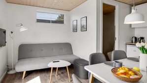 Un salon moderne avec un canapé gris, une petite table ronde, une table à manger agrémentée d’un panier de fruits, et un coin cuisine équipé d’une bouilloire et d’une machine à café.