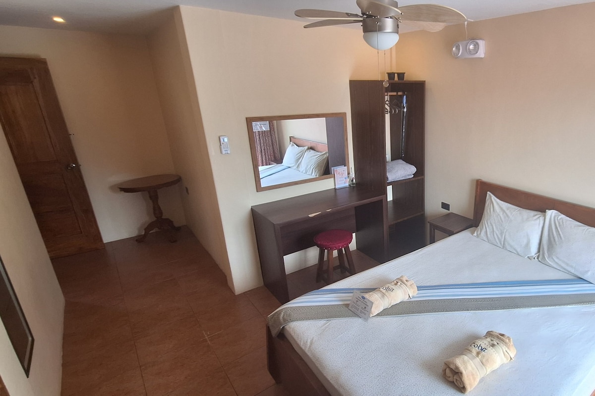 A hotel room with a bed, a desk, a chair, a mirror, a ceiling fan, and a door.